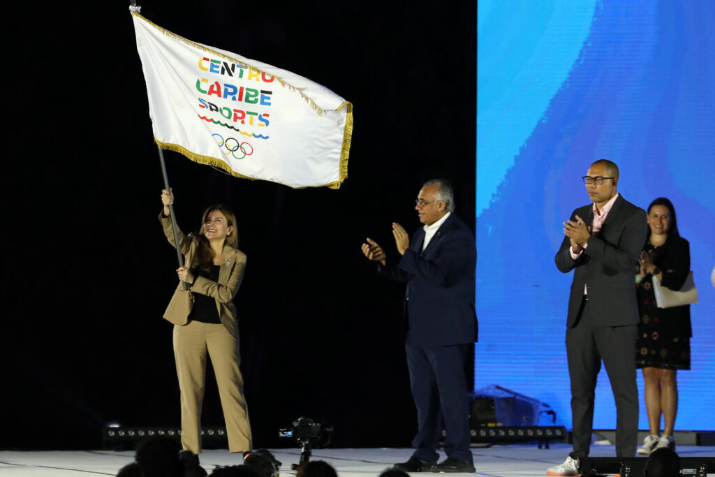 La alcaldesa de Santo Domingo Carolina Mejía ondea la bandera de Centro Caribe Sports junto al presidente de Centro Caribe Sports Luis Mejía Oviedo y el presidente de Comité Organizador de los Juegos Centroamericanos y del Caribe en San Salvador, Yamil Bukele, en la clausura en los Juegos Centroamericanos y del Caribe, en San Salvador.